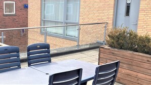 Een terras buiten met een tafel en stoelen, een houten plantenbak, en een gebouw met een blauwe deur en ramen.