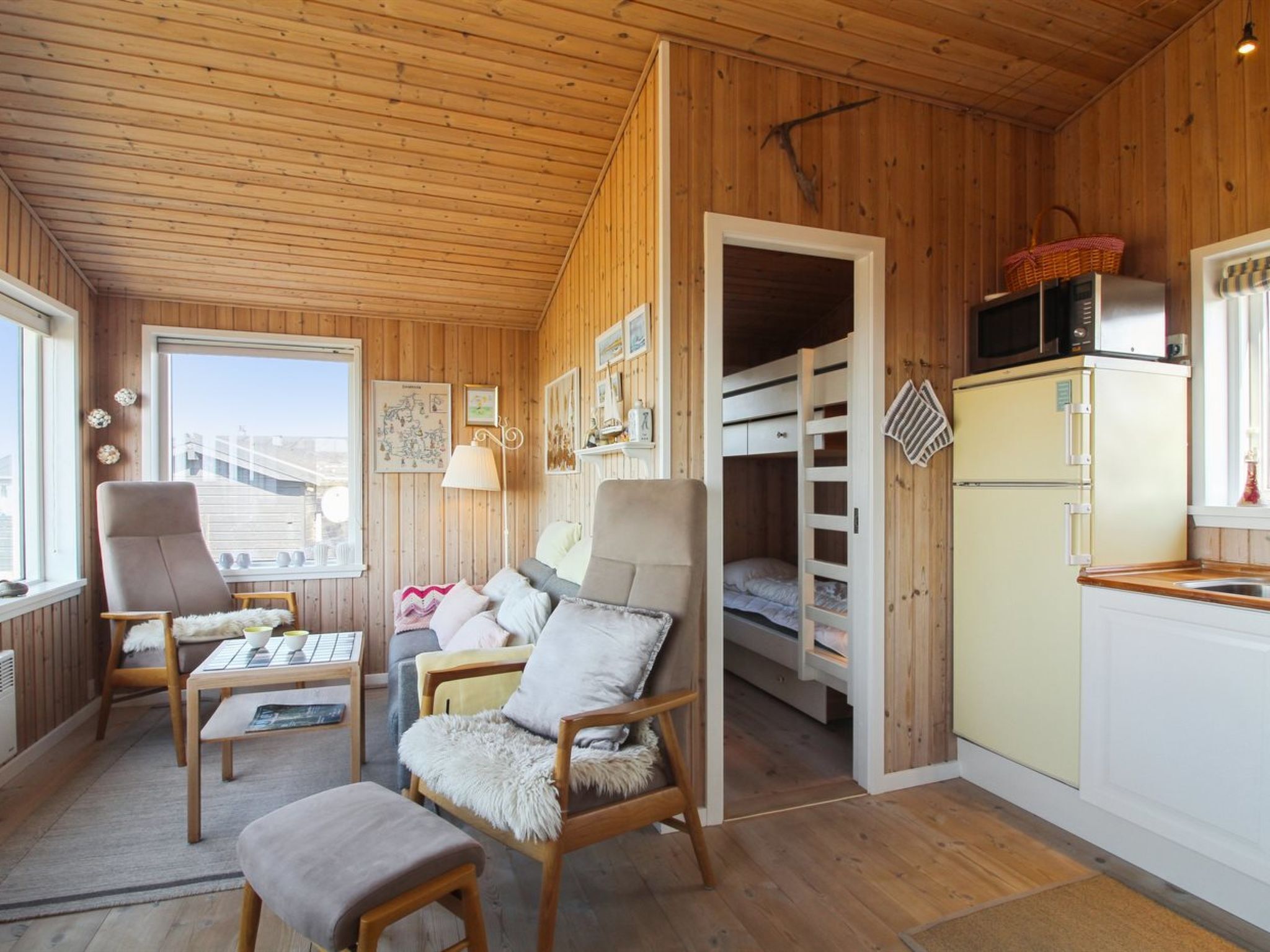 A cozy cabin interior with wooden walls, a dining area, a living room with a sofa and chairs, a kitchen with a refrigerator, and a window with a view.