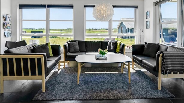 Een moderne woonkamer met een zwarte leren zithoek, een witte salontafel en een groot raam met uitzicht op het landschap.
