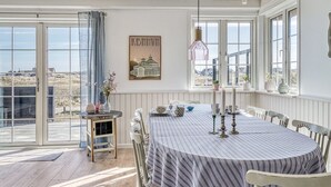 A dining area with a round table, chairs, a vase with flowers, a candle holder, and a view of the landscape through large windows.
