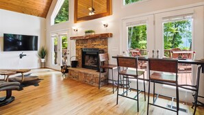 A spacious living room with a stone fireplace, wooden flooring, and large windows offering a view of greenery. - 16 mi to DT, Hot Tub, Fire Pit, FP, Basket Ball Court, Mt View, Secluded & Quiet (Barnardsville)
