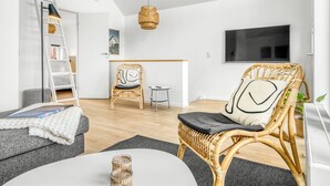 Een moderne woonkamer met een bank, een rieten fauteuil, een klein tafeltje, een ladder, een hanglamp en een televisie aan de muur.