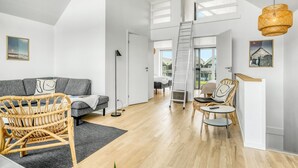 Een ruime woonkamer met een grijze bank, rieten stoelen, een kleine tafel en een ladder naar een hoger niveau.