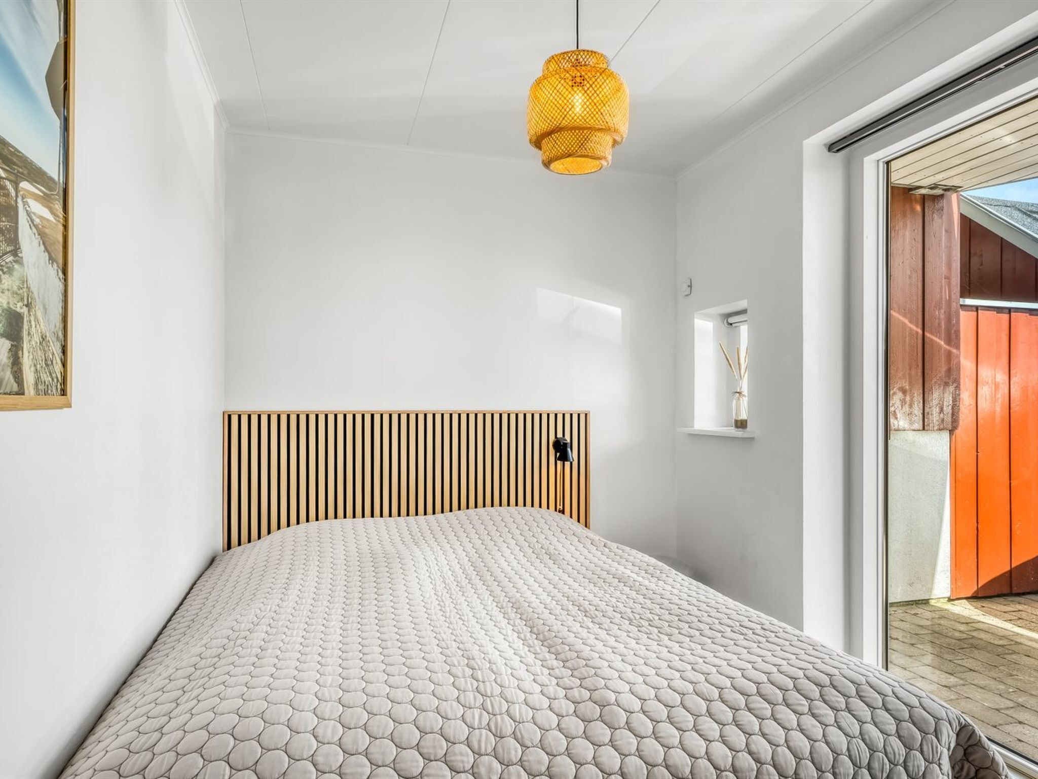 A bedroom with a bed, a wooden slat headboard, a hanging light fixture, a small window, and a view of the outside.