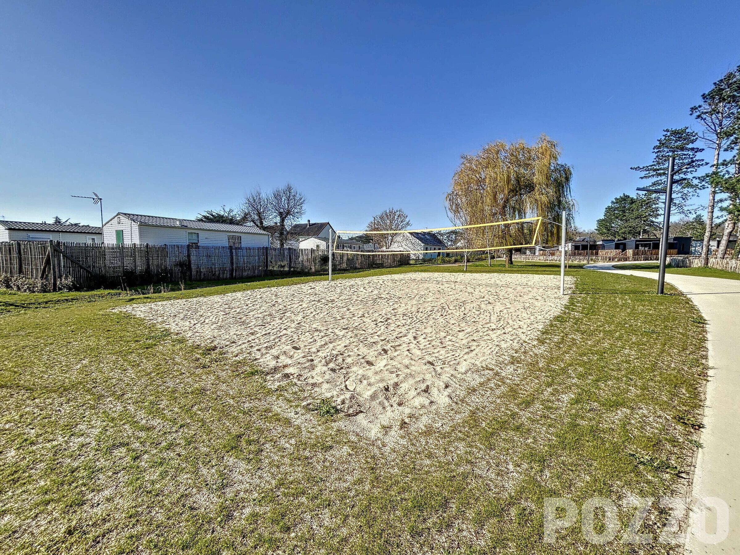 A sandy volleyball court with a net, surrounded by a wooden fence and trees.
