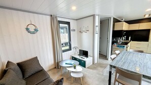 Kompakti olohuone, jossa on sohva, pieni pyöreä pöytä, televisiotaso ja sen päällä televisio sekä keittiöalue, jossa on puiset kaapit ja työtaso.