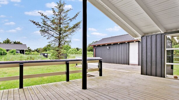 Une terrasse en bois couverte, dotée d’une rambarde en métal, un grand arbre et un bâtiment à l’extérieur sombre et au toit recouvert de tuiles.