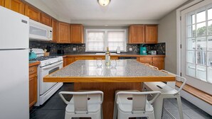 Une cuisine avec des meubles en bois, un îlot central doté d’un plan de travail en granit, des chaises blanches et une vue dégagée grâce à de grandes fenêtres.