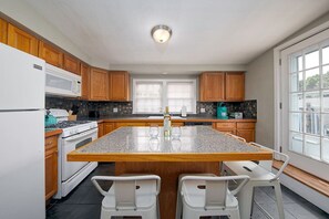 Una cocina con gabinetes de madera, una isla con encimera de granito, sillas blancas y vistas amplias a través de grandes ventanales.