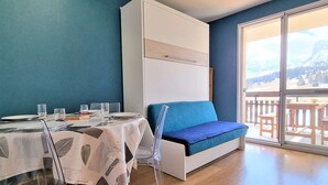 Une pièce comprenant une table à manger dressée pour quatre personnes, un canapé bleu et une grande fenêtre donnant sur un paysage montagneux.