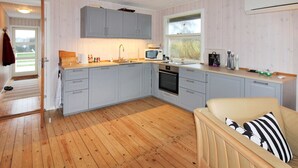 Cocina compacta con gabinetes grises, un microondas y un lavamanos. El suelo es de madera y hay un sofá color crema con almohadones a rayas.