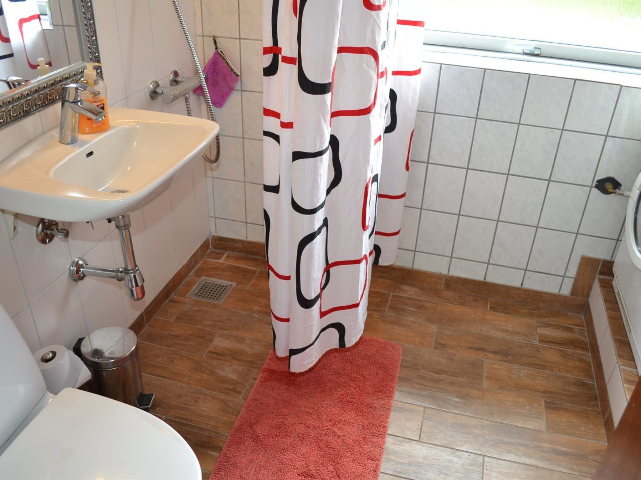 A bathroom with a white sink, a mirror, a shower curtain with a geometric pattern, a red bath mat, and tiled flooring.