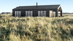 Un edificio moderno de una sola planta, con techo inclinado, ventanas amplias y chimenea. Se encuentra en un campo cubierto de hierba bajo un cielo despejado.
