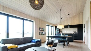 Una sala de estar moderna con sofá, mesa de centro y mecedora, un área de cocina con mesa de comedor, y ventanas amplias que dejan entrar luz natural.
