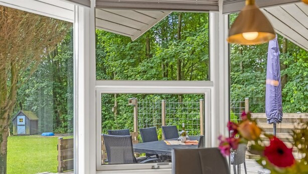 En overdækket terrasse med bord og stole, omgivet af glasvægge der giver udsigt til en frodig have og et mindre udhus.