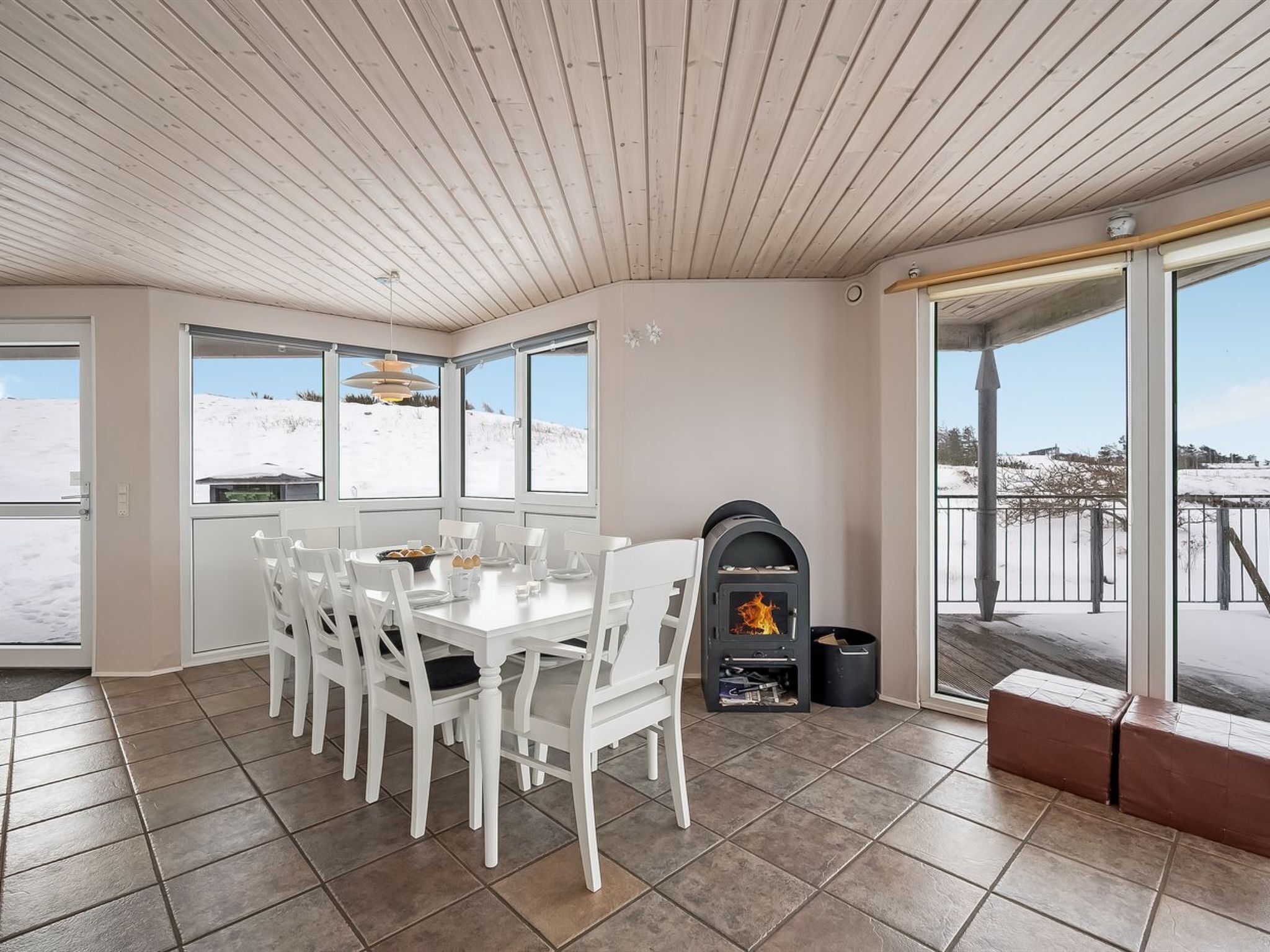 Un coin repas avec une table et des chaises blanches, un poêle à bois et de grandes fenêtres donnant sur un paysage enneigé.