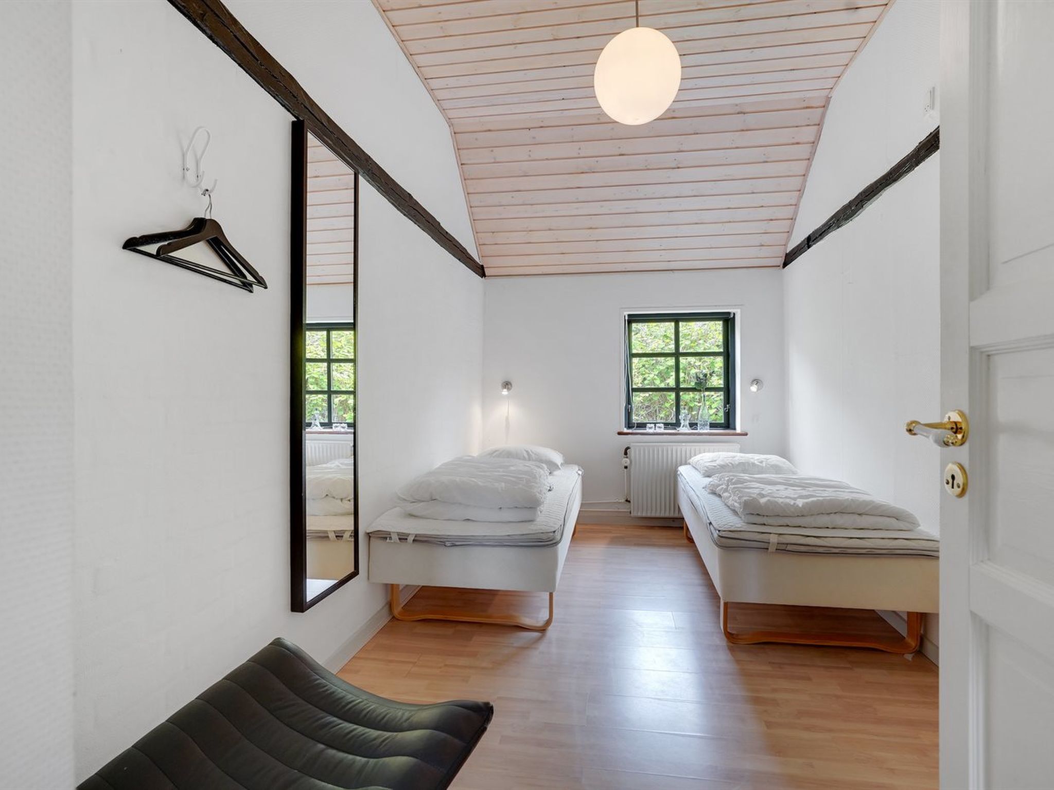 A bedroom with two beds, a hanging light fixture, a radiator, a window, and a mirror on the wall.
