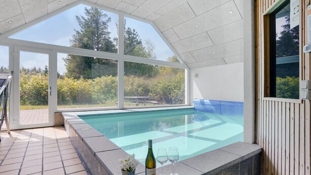 A modern indoor swimming pool with clear glass walls, a stone pool edge, and a wooden deck area.
