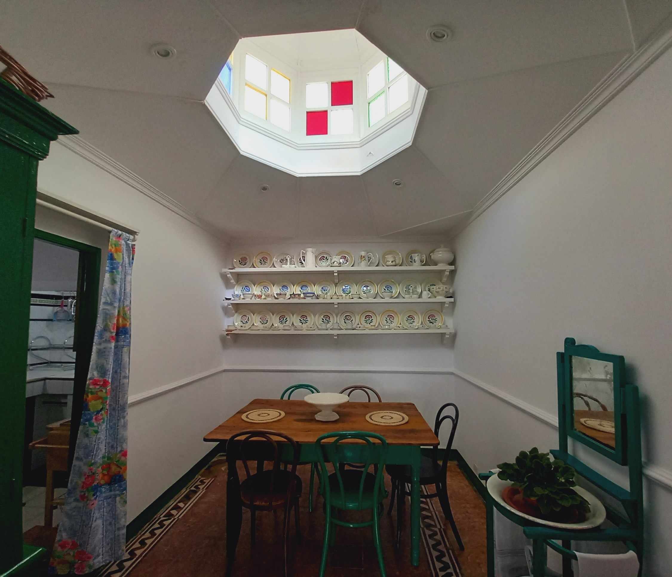 A dining area with a wooden table, green chairs, a ceiling skylight, and shelves with dishes.