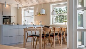 A modern kitchen with wooden cabinets, a dining area with wooden chairs, pendant lights, and large windows offering natural light.