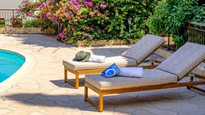 Uma área à beira da piscina com duas espreguiçadeiras de madeira, uma delas com um chapéu e um livro, e piso de ladrilho de pedra.