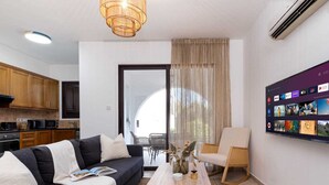 Uma sala de estar moderna com um sofá cinza escuro, uma mesa de centro de madeira clara e uma poltrona branca. Uma televisão de tela plana grande está instalada na parede, exibindo diversos canais.