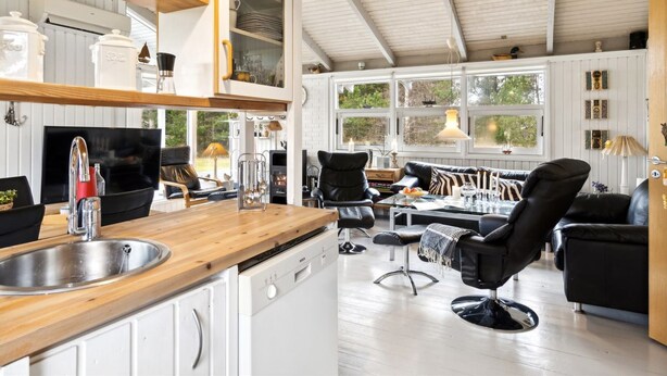 A modern kitchen with white cabinetry, a wooden countertop, and a built-in dishwasher. There is a black leather sofa and a dining table with chairs in the adjacent living area. Large windows allow natural light to fill the space. - "Alvine" - 475m from the sea by Interhome (Jerup)