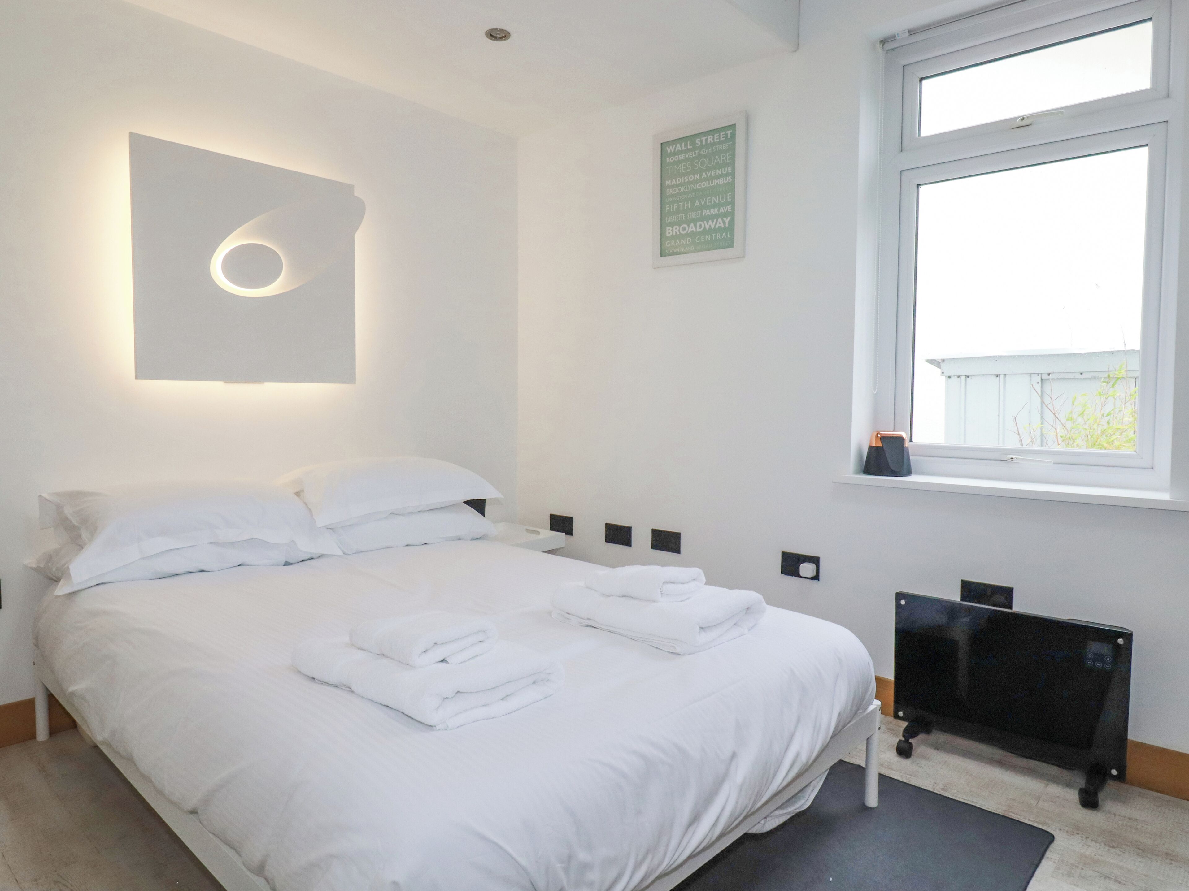 A neatly made bed with white linens and towels, a wall-mounted light fixture, a framed sign with street names, and a window with a view of the outside.