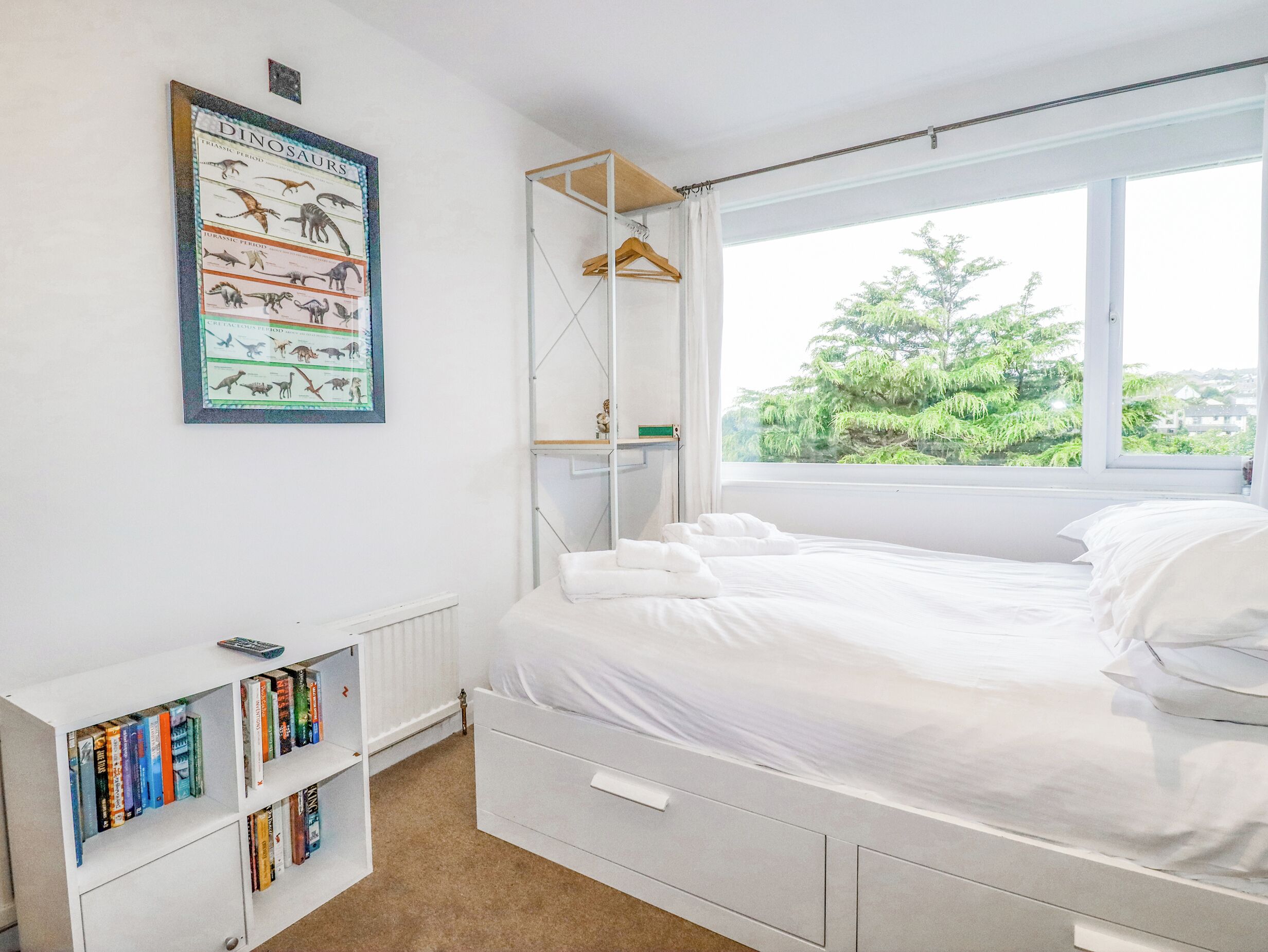 A bedroom with a bed, a bookshelf, a radiator, and a window with a view of greenery.