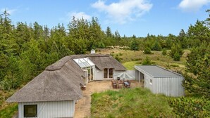 Une maison à toit de chaume avec un patio, un bâtiment séparé et une colline boisée en arrière-plan.