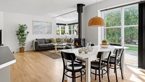 Un salon moderne avec une cheminée, un coin repas avec une table blanche et des chaises noires, et une grande fenêtre donnant sur un paysage verdoyant.