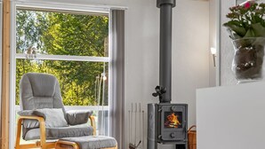 A modern living room with a grey upholstered chair, a black wood-burning stove, and a large window with a view of trees. - "Barne" - 4km from the sea by Interhome (Sæby)