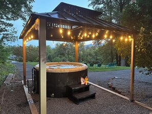 Un bain à remous installé sous une pergola en bois, illuminé de guirlandes lumineuses, et entouré de gravier et de verdure.