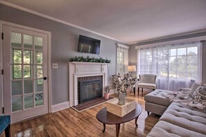 A living room with a fireplace, a sofa, a television mounted above the fireplace, and a large window with sheer curtains.