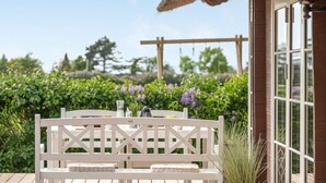 A patio with a white wooden table and bench, a thatched roof, and a view of a garden with purple flowers.