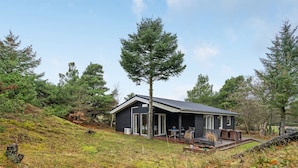 A cabin with a deck and outdoor seating, surrounded by trees and a grassy hill.