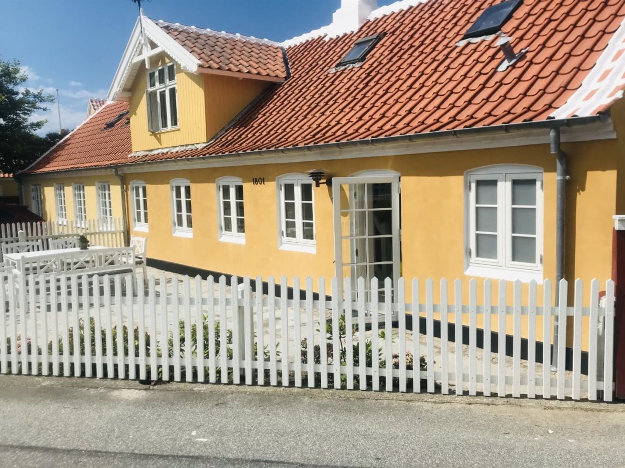 Un edificio amarillo con techo de tejas rojas, ventanas blancas y valla de piquetes blanca.