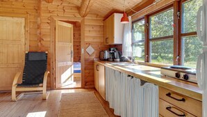 A wooden cabin interior with a kitchen area, a dining chair, and a window with a view of trees.