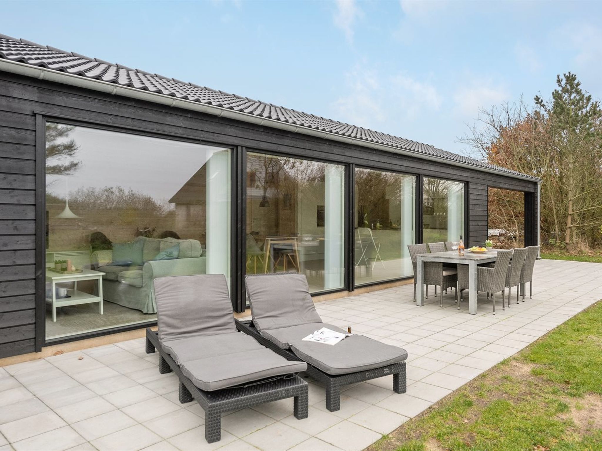 Een modern huis met grote glasramen, een patio met twee loungesteunen en een tafel, en een tuin.