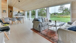 Ein modernes Interieur mit Holzboden, einem Essbereich, einem Wohnzimmer mit Sofa und Couchtisch sowie großen Fenstern mit Blick ins Freie.