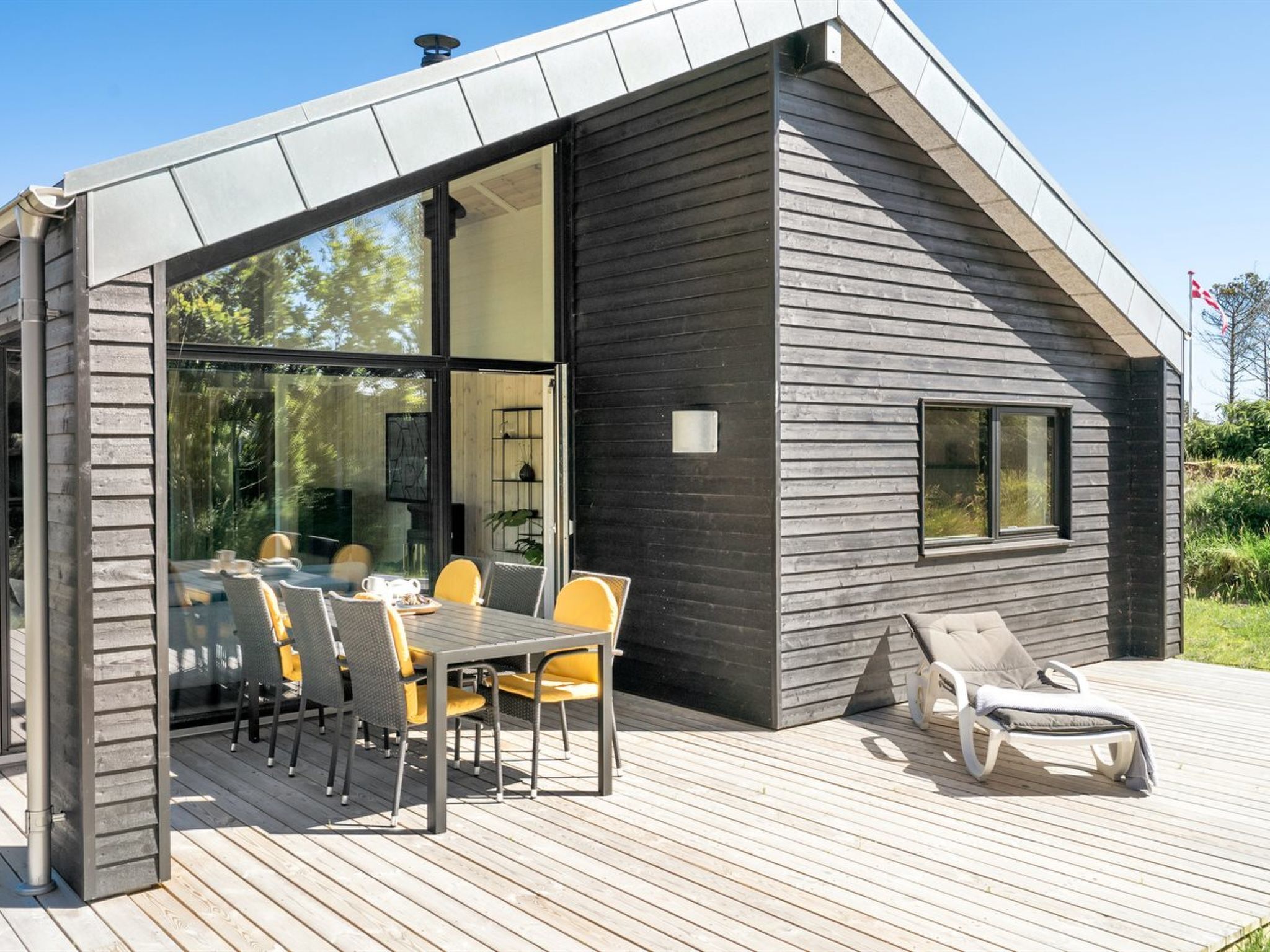 Ein modernes Haus mit einer Terrasse, einem Essbereich mit Stühlen und einem Tisch sowie einem Liegestuhl.