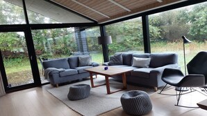 A modern living room with a large glass wall, a wooden ceiling, and a grey sofa set.