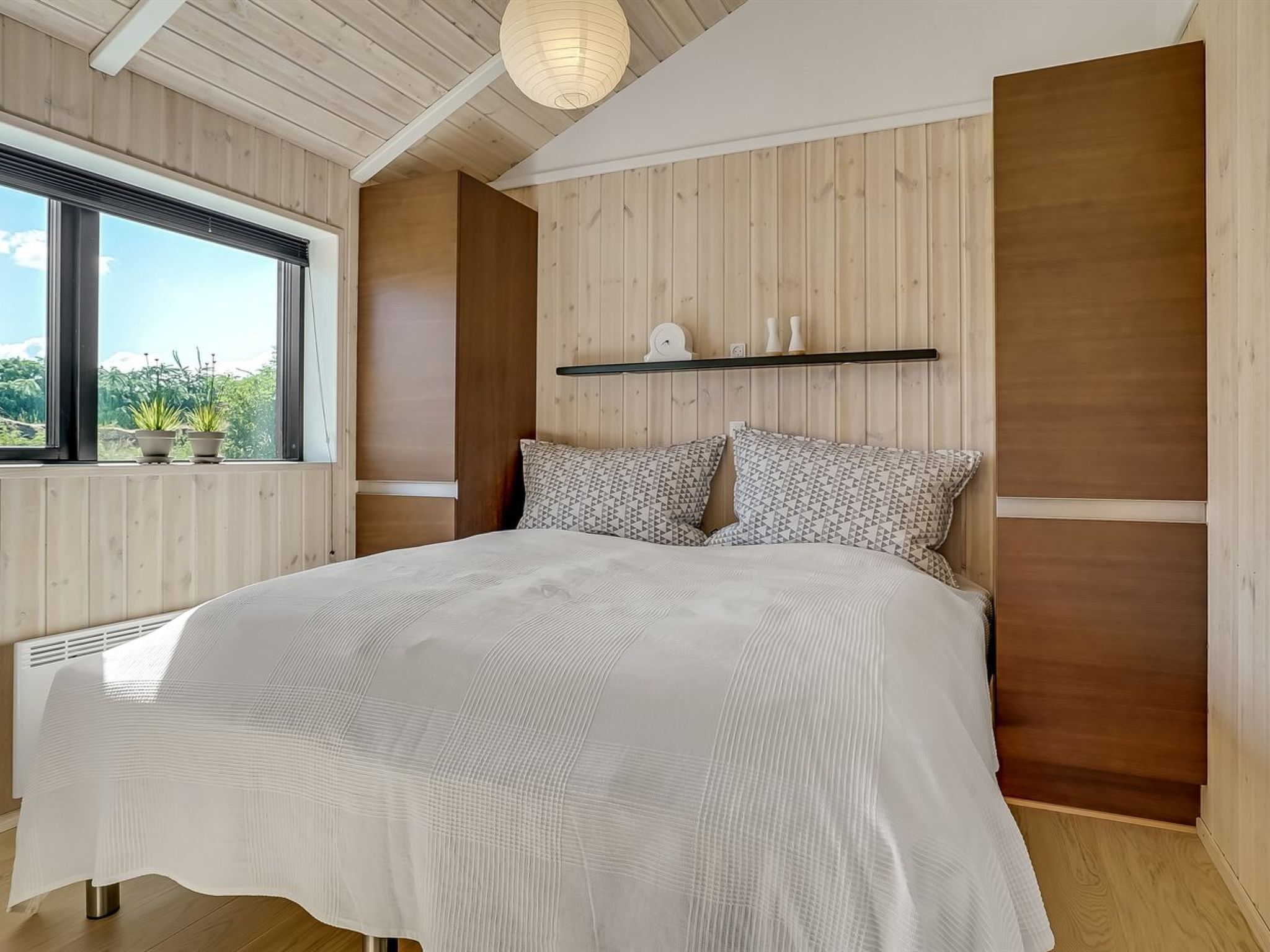 Een slaapkamer met een groot bed, houten paneelwanden, een raam met planten en een plafondlamp.
