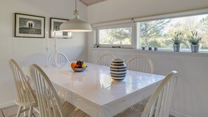A dining area with a white table, white chairs, a hanging light fixture, and a window with potted plants.