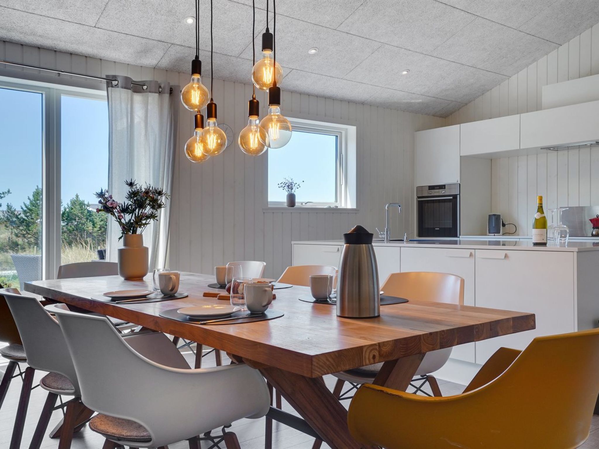 Une cuisine moderne avec une table à manger en bois, des chaises blanches et jaunes, et des luminaires suspendus.