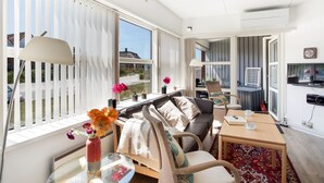 A modern living room with a sofa, armchairs, a glass coffee table, and a dining table. There are large windows with blinds, a view of the outside, and a small balcony with a chair.