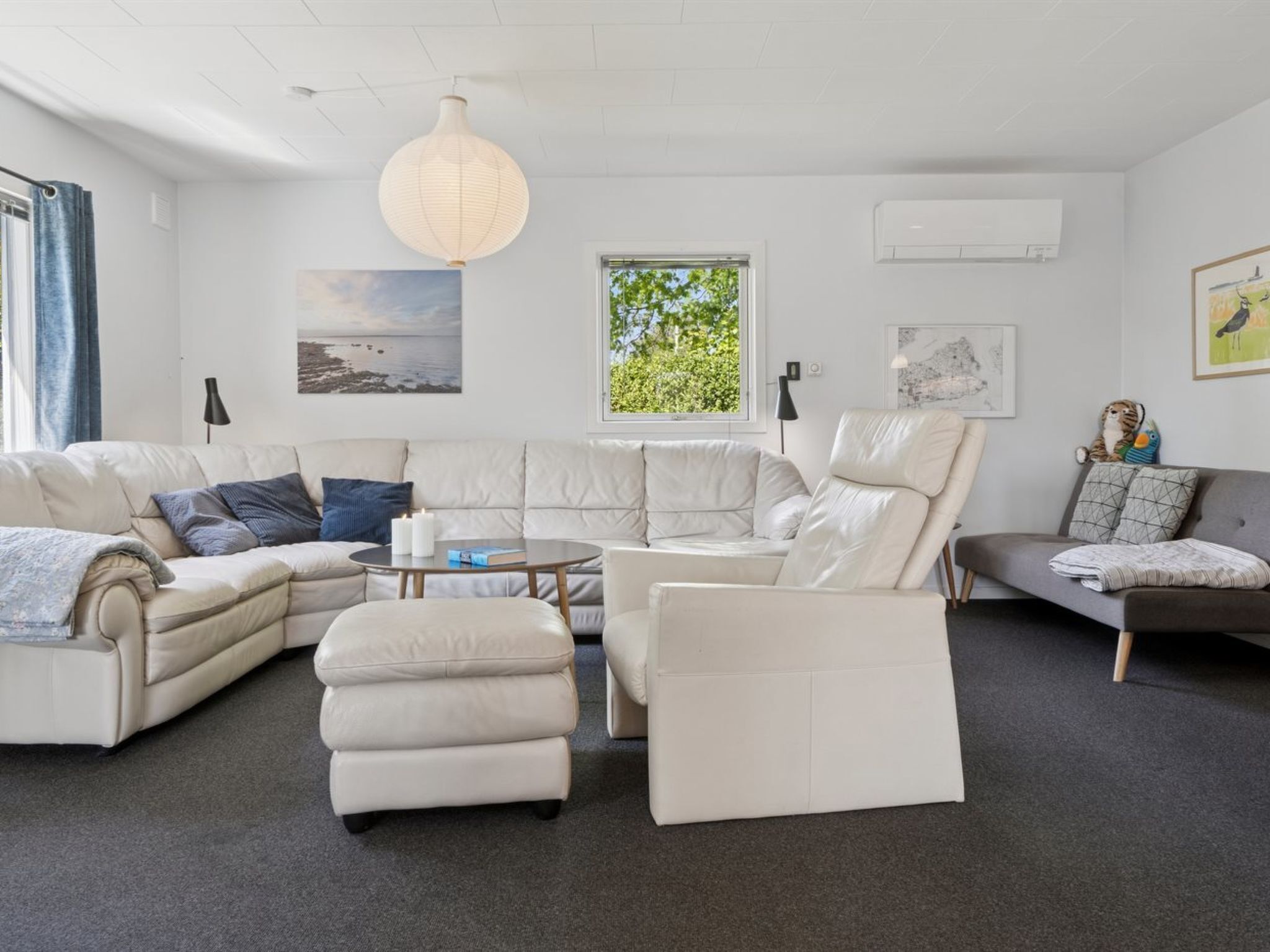 A modern living room with a sectional sofa, a round coffee table, and a small ottoman. There is a large window with a view of trees, a hanging light fixture, and artwork on the walls.
