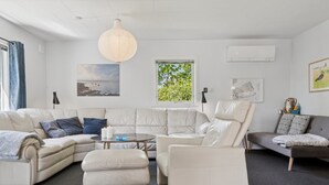 A modern living room with a sectional sofa, a round coffee table, and a small ottoman. There is a large window with a view of trees, a hanging light fixture, and artwork on the walls. - "Ømund" - 600m to the fjord by Interhome (Holbæk)