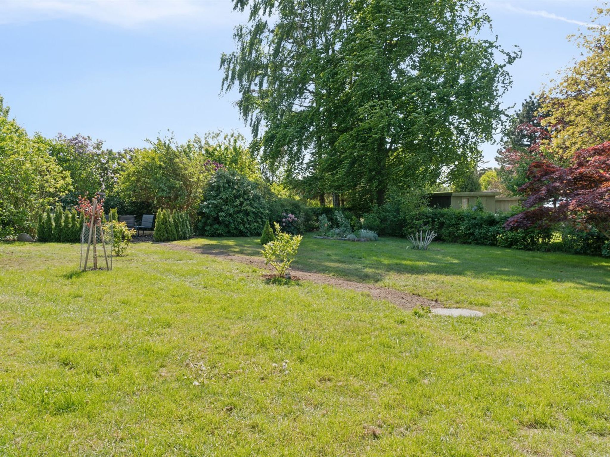 A well-maintained garden with a variety of trees and shrubs, a small wooden structure, and a clear sky.