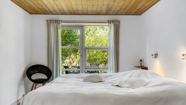 Un dormitorio con una cama grande, una ventana con cortinas, una silla y una mesita de noche con una lámpara.